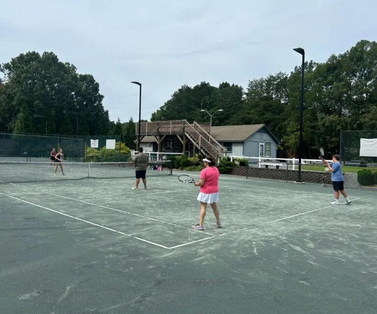 Adults playing doubles tennis with the clubhouse in the background at Asheboro Racquet & Swim Club
